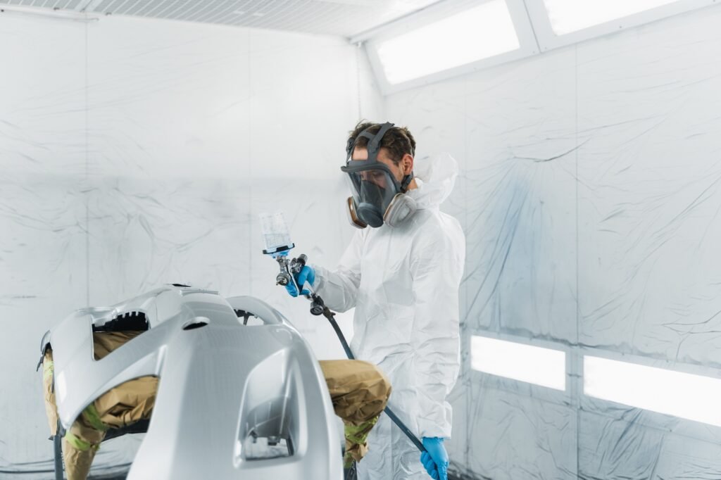 Car painter in a protective suit and mask varnishes a painted bumper of a vehicle