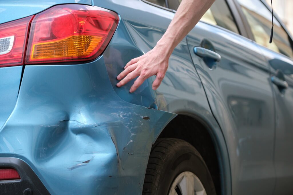 Driver hand examining dented car with damaged fender parked on city street side.