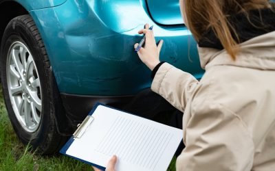 Person inspecting car bumper damage with clipboard outdoors.