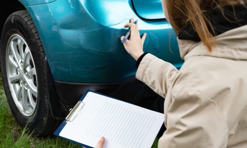 Person inspecting car bumper damage with clipboard outdoors.